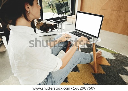 Stylish hipster girl using laptop with blank screen, sitting on floor  in modern room. Young woman shopping or working online from home mock up. Freelance and freelancer