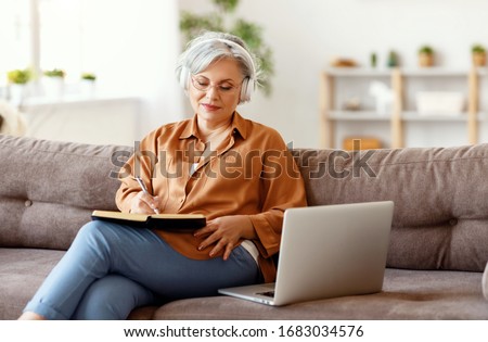 Elderly female in headphones and glasses writing in notebook and listening to music while sitting on couch near laptop
 Royalty-Free Stock Photo #1683034576