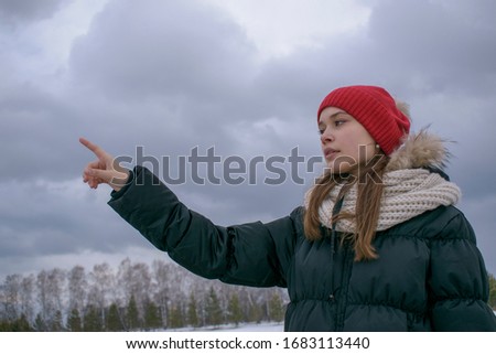 Portrait of a girl pushing a virtual button. Profile view. Winter, warm clothes, red hat, white snow. Background forest, sky with clouds. Touch control concept.