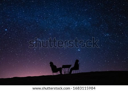 young couple sitting at a table on a desert dune while talking, relaxing and observing the stars and the milky way above them Royalty-Free Stock Photo #1683115984