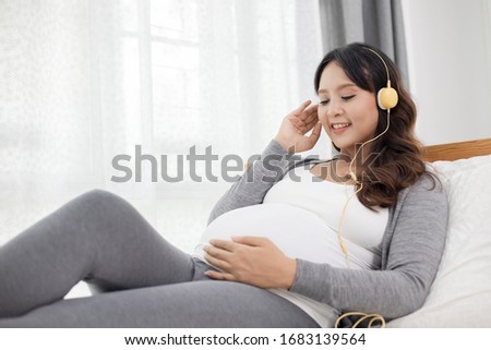 Happy Asian pregnant woman sitting and listening music on bed at home bedroom, pregnancy, People and maternity concept