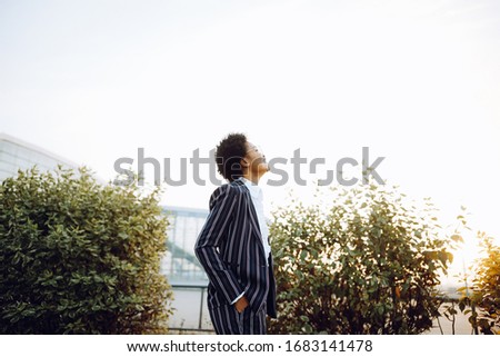 4k. Travel, Digital. A charming African American woman in an elegant striped suit, looks at the sky, smiles and feels fantastic. She stands on background of bushes near the airport. Businesswoman or