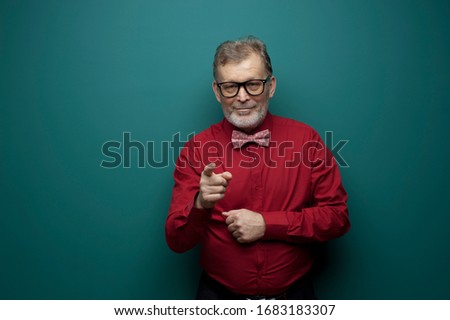 Portrait of a positive elderly man in glasses with bow-tie and red shirt posing on a gray-green background. Concept of positive successful retiree. Copyspace