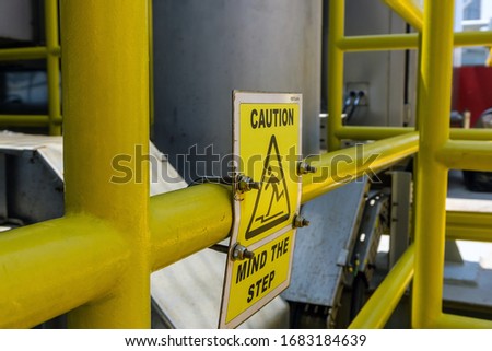 Signage with icon and letter 'Caution Mind The Step' on a handrail on board a oil production platform
