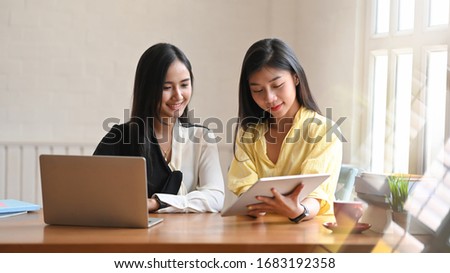 Photo of young women talking together while sitting at the wooden working table with computer laptop and tablet next to living room windows as background.