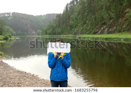 young man hides his face behind white banner with copy space. model is photographed standing on banks of river