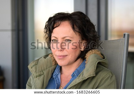 Attractive thoughtful serious brunette woman with curly hair seated in a chair on an outdoor patio looking aside with a faraway dreamy expression and quiet smile