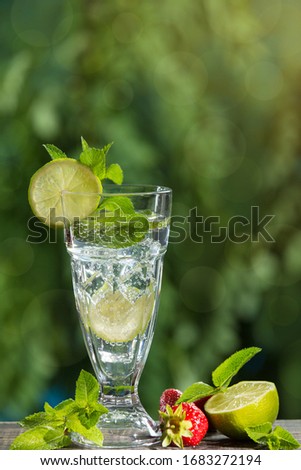 in a tall glass a refreshing drink with the addition of mint, lime and ice, against a background of greenery, next to strawberries, lime and mint leaves