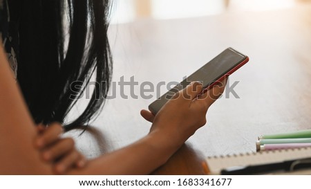 Cropped image of young creative woman's hands holding a smartphone with empty screen while sitting at the working desk over comfortable living room as background.