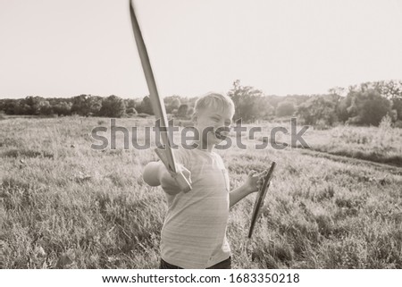 Happy white kid playing outdoor with wooden toys standing in beautiful summer sunset countryside landscape. Boy holding sword and shield in hands happily. 