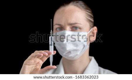 A syringe with a medicine injection in the hands of female doctor in white medical gown with sterile face mask on black background. Anti virus human health concept. Medicine and vaccination. 