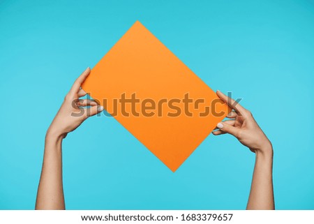 Studio photo of elegant woman's hands with white manicure holding orange squared paper inclined while standing over blue background. Human gesturing concept