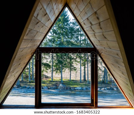 Log cabin window. Looking through the window from inside. Nature behind window glass. Window frame and curtain. Triangle. Timber construction. Wooden, log hut. Warm wooden shack. Sunny weather Royalty-Free Stock Photo #1683405322