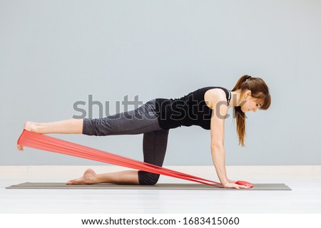 Sideways of sporty European woman has workout with rubber red band, dressed in black sportsclothes, has exercises for buttocks, poses on knees at yoga mat against grey background. Motivation concept.