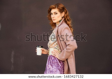 Photo of a fashionable beautiful woman with long hair in the spring city in the sunshine outdoors with a cup of coffee in her hands on the background of a building wall