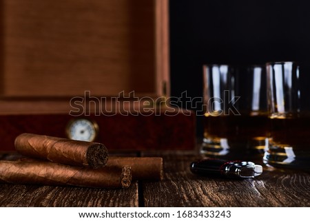 Still life with three cuban cigars, two glasses of whiskey or rum, lighter and wooden box with hygrometer. Old wooden table top and black background. Space for your text. Royalty-Free Stock Photo #1683433243