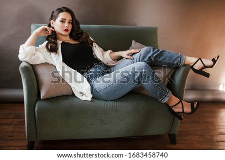 a brunette girl with curly hair with a make up red lips in a black t-shirt white blouse jeans sandals with heels in a photo Studio indoors at home in an apartment smiling posing laying on the sofa