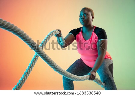 Young african-american plus size female model's training on gradient background in neon light. Doing workout exercises with the ropes. Concept of sport, healthy lifestyle, body positive, equality.