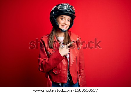 Young beautiful brunette motocyclist woman wearing motorcycle helmet and red jacket cheerful with a smile of face pointing with hand and finger up to the side with happy and natural expression on face