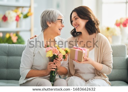 Happy mother's day! Beautiful young woman and her mother with flowers and gift box at home.                                 Royalty-Free Stock Photo #1683662632