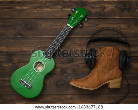 A ukulele and a suede boot with earbuds on a wooden background. Retro technique for playing music.