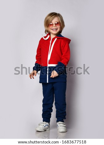 Happy smiling laughing blond kid boy in red and blue tracksuit sportwear and aviator sunglasses poses over white background