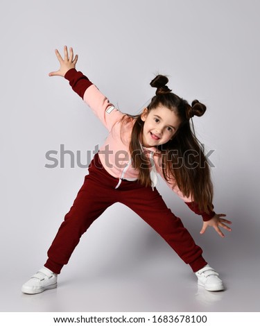 Frolic curious kid girl with straight brunette hair with buns in modern fashion pink brown sportwear bending over with her arms outstretched and legs wide apart over gray background
