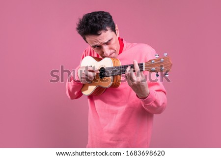 Serious concentrated guy with ukulele in hands playing alone and looking serious. Aiming with instrument. Isolated over pink background