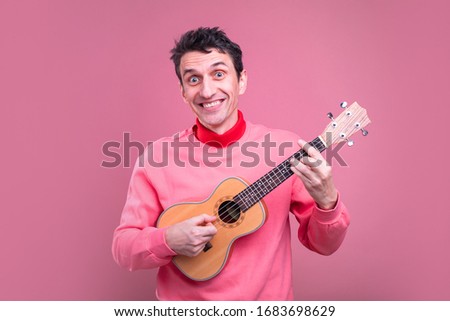 Picture of classical guitarist professional with ukulele in hands playing and have fun alone. Smile on camera. Isolated on pink background