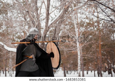 Viking in winter with a spear and a round shield of red-black color. A guy in a helmet and chain mail in the snow. A man in armor goes to battle