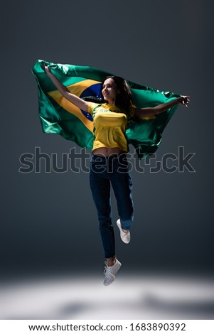 attractive cheerful football fan jumping with brazilian flag on grey