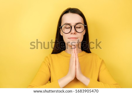 Calm young woman practicing yoga, breathing, mindful young female meditating with closed eyes, namaste gesture, healthy lifestyle stress relief concept, wears jumper, isolated on yellow background