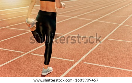 Woman in sportswear before a run makes a stretch holding her leg with her hand on a stadium track with a red coating. Crop photo