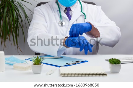 doctor in a white medical coat sits at a table in a brown leather chair and puts on sterile medical gloves in his hands before receiving and examining patients