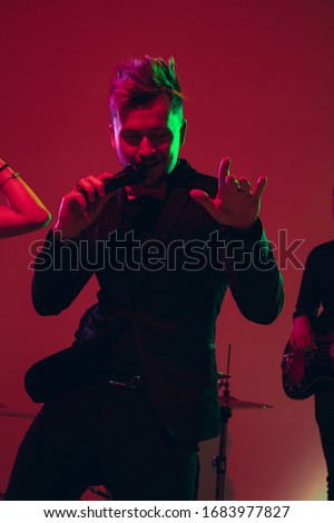 Young and joyful caucasian singer performing on red background in neon light. Concept of music, hobby, festival. Colorful portrait of modern artist. Attented, inspired. Art, cover band. Close up.