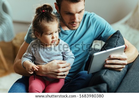 Happy father and his small daughter watching cartoons on touchpad while relaxing at home. 