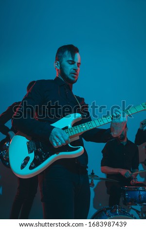 Young caucasian musicians, band performing on blue studio background in neon light. Concept of music, hobby, festival. Colorful portrait of modern artist. Attented and inspired. Art, cover band.