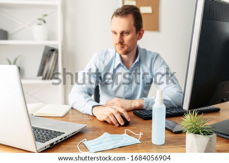 Precautions to prevent the spread of viral diseases, coronavirus, covid 19. Mask and disinfectant on the desktop, a man with a laptop works on background. Keep yourself safe