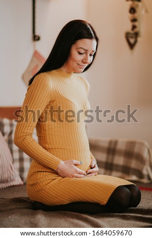Pregnant young woman touching baby bump. Young woman expecting baby kneeling on bed touching tummy and smiling.