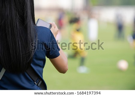 Mother standing, watching and taking pictures of her daughter playing football in a school tournament on a clear sky and sunny day. Sport, active lifestyle, happy family and soccer mom concept.