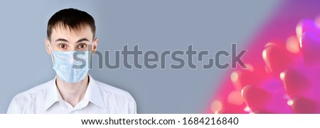 Man in a medical mask. Surprised, scared man  in a white shirt looking at camera, on a gray background, on the right is a large red Coronavirus bacterium. Banner 