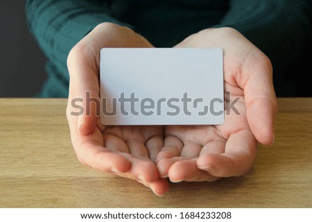 A white rectangular card lies in the opened palms of a girl in a blue sweater. Mockup for design, place for text, copy space.