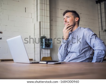 thinking remote online working man having his hand on chin and looking up in casual outfit with laptop and notebook sitting in an coworking office at a work desk