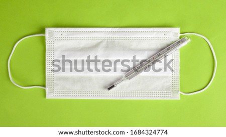 medical face mask with thermometer on a light green background top view. Coronavirus flu protection and screening, quarantine pandemic covid-19.