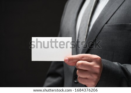 white blank business card closeup in businessman hand, gray suit, dark wall background