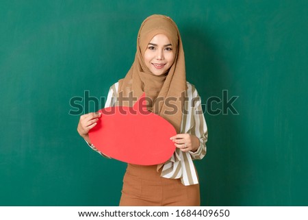 Young and beautiful Asian Muslim woman standing at green wall,  holding and showing colorful blank speech bubble. Concept for speak out to communicate important text of messeges to social or public