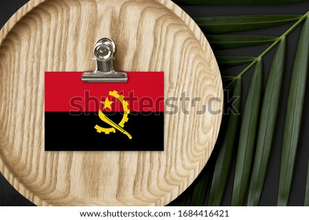 Angola flag tagged on wooden plate. Tropical palm leaves monstera on background. Minimal national concept.