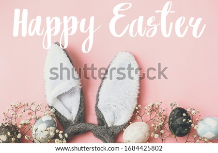 Happy Easter text. Easter greetings card. Stylish easter eggs, bunny ears, flowers border flat lay on pink wallpaper. Natural dyed marble easter egg and grey rabbit ears. Holiday card