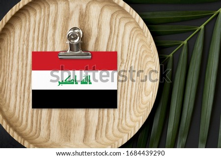 Iraq flag tagged on wooden plate. Tropical palm leaves monstera on background. Minimal national concept.