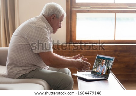Side view middle aged mature older man sitting on sofa, looking at computer screen, holding video call listening grown up handsome son, explaining how important stay at home during virus outbreak. Royalty-Free Stock Photo #1684479148